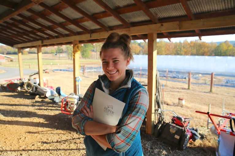 Young female farmer holding a Heifer packet and smiling while standing in an enclosed shelter.