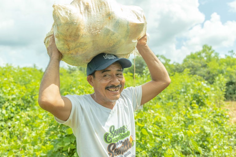 A smiling man in a white t-shirt and cap carries a large sack of squash rinds above his head.